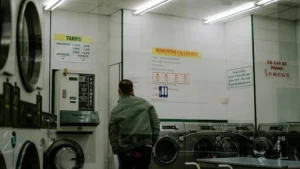 Man in a laundromat inspecting commercial laundry machines, highlighting the importance of proper maintenance to extend their lifespan & ensure efficiency.