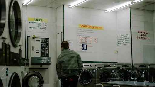 Man in a laundromat inspecting commercial laundry machines, highlighting the importance of proper maintenance to extend their lifespan & ensure efficiency.