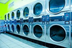 Row of industrial stainless steel dryers in a commercial laundromat, illustrating high-capacity smart laundry equipment for faster turnaround.